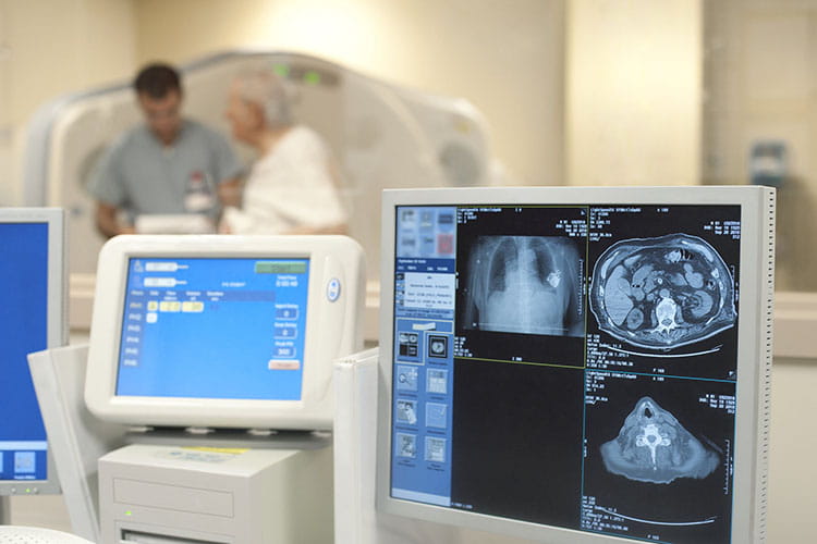 Close-up of diagnostic imaging screens with a male techinican and an elderly male patient standing in front of the equipment in the background.