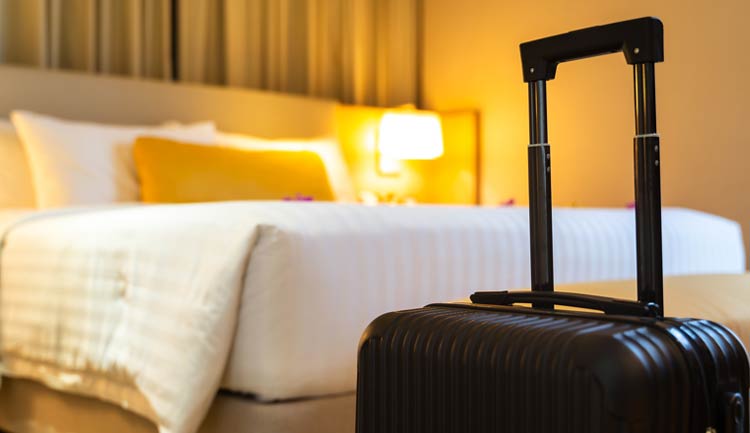 A wheelie suitcase sitting on he floor in front a bed in a hotel room.